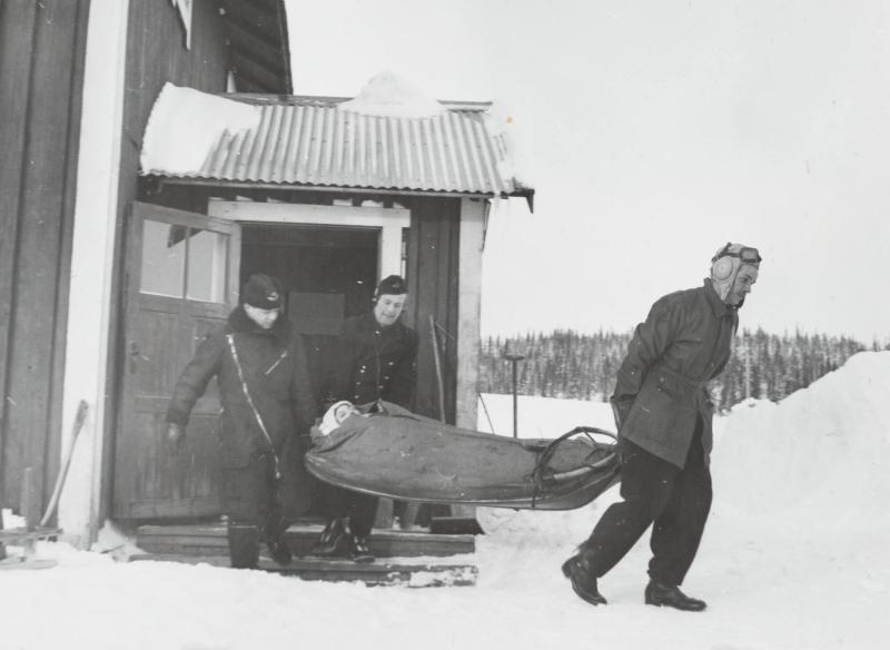 The ambulance plane has landed on a lake at some distance from the farm and the pilot with the aid of two attendants are carrying the patient out of the house